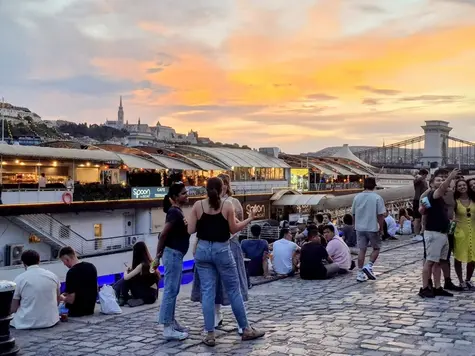 Turisti e locali si rilassano sul lungofiume del Danubio a Pest vicino al battello Spoon al tramonto con il Bastione dei Pescatori sullo sfondo