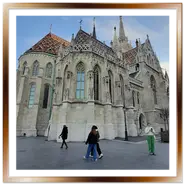 Vista posteriore dell'abside gotica della Chiesa di Mattia a Budapest, con i dettagli delle finestre ad arco a sesto acuto, il tetto decorato e i passanti sulla piazza pedonale pavimentata sotto un cielo parzialmente nuvoloso.<br />