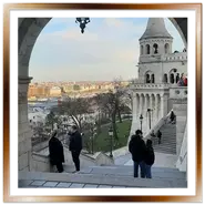 Vista panoramica attraverso un arco in pietra del Bastione dei Pescatori a Budapest, che mostra una torre conica bianca, le scale esterne con turisti e lo skyline di Pest sullo sfondo.