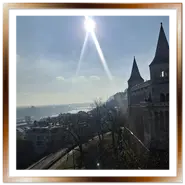 Veduta panoramica invernale dal Bastione dei Pescatori a Budapest, con le torri fiabesche sulla destra, il sole splendente che crea un riflesso a stella nel cielo chiaro e la città nebbiosa lungo il Danubio sullo sfondo.