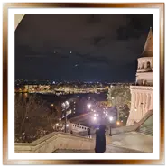 Vista notturna di Budapest e del fiume Danubio illuminati, incorniciata da un arco in pietra e dalle scale del Bastione dei Pescatori sulla collina di Buda.