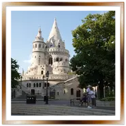 Le torri bianche in stile neogotico e neoromanico del Bastione dei Pescatori a Budapest viste dalla piazza, circondate da alberi verdi con alcuni turisti che si riposano sulle panchine e scattano foto.