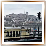 Panorama del Castello di Buda e del Palazzo Reale con lievi chiazze di neve, visto dal lungodanubio di Pest, con la biglietteria Dunacorso e i binari del tram in primo piano sotto un cielo nuvoloso.