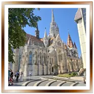 La Chiesa di Mattia vista dalla piazza vicino al Bastione dei Pescatori a Budapest, con la pavimentazione a cerchi concentrici in primo piano, turisti e il tetto in maiolica colorata sotto un cielo sereno.