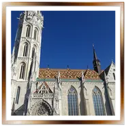 Vista esterna dell'architettura gotica della Chiesa di Mattia (Matthias Church) a Budapest, Ungheria. Si vedono la grande torre campanaria in pietra bianca, il tetto con le tegole Zsolnay colorate e i turisti vicino all'ingresso principale sotto un cielo azzurro e sereno.
