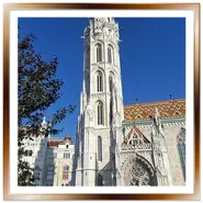 Inquadratura laterale della Chiesa di Mattia a Budapest durante un tour di Buda, con la torre campanaria gotica in pietra bianca in evidenza, il portale d'ingresso con rosone, turisti sulla piazza e il tetto colorato sotto un cielo blu.