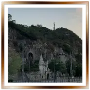 Vista esterna della Chiesa nella Roccia (Sziklatemplom) scavata nella collina Gellért a Budapest. Si vedono l'ingresso della grotta, le torrette in pietra, la grande croce sulla cima e la Statua della Libertà sullo sfondo al tramonto.