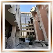 Prospettiva verticale del cortile dei Domenicani a Budapest, con le antiche mura gotiche color mattone integrate tra le moderne vetrate riflettenti dell'hotel Hilton e un piccolo albero al centro.