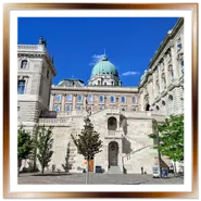 Vista del cortile interno del Palazzo Reale nel Castello di Buda a Budapest, con l'imponente scalinata in pietra bianca, la cupola verde dell'edificio principale sullo sfondo e alberi decorativi sotto un cielo blu limpido.