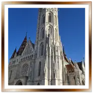 Inquadratura frontale dal basso della Chiesa di Mattia a Budapest, con la grande torre campanaria neogotica in pietra bianca, il rosone della facciata e i dettagli del tetto colorato sotto un cielo blu limpido.