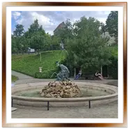 La Fontana del Tritone circolare in pietra nei Giardini Reali del Várkert Bazár a Budapest, con zampilli d'acqua, alberi ed edera sullo sfondo e persone sedute su una panchina.