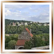 Antica torre fortificata in pietra con tetto in tegole rosse e mura del Castello di Buda in primo piano, con vista sul verdeggiante monte Gellért punteggiato da abitazioni storiche sotto un cielo nuvoloso.