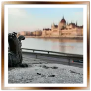 Inquadratura ravvicinata di una mini statua in bronzo di Kolodko sul muretto del lungofiume di Buda, con il fiume Danubio e il Palazzo del Parlamento di Budapest sfocati sullo sfondo al tramonto.