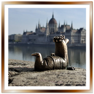 La mini statua in bronzo del verme Főkukac dell'artista Kolodko sul lungofiume di Buda, con il Palazzo del Parlamento di Budapest sfocato sullo sfondo.