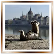 La mini statua in bronzo del verme Főkukac dell'artista Kolodko sul lungofiume di Buda, con il Palazzo del Parlamento di Budapest sfocato sullo sfondo.