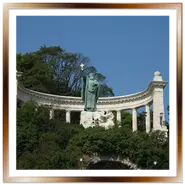La grande statua in bronzo verde di San Gerardo che solleva una croce, situata davanti a un colonnato bianco neoclassico sul fianco boscoso del monte Gellért a Budapest, sotto un cielo azzurro limpido.