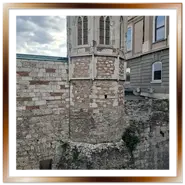 Veduta ravvicinata delle antiche mura in pietra del Castello di Buda a Budapest. Si notano i diversi strati di costruzione, le feritoie e le parti più rovinate che testimoniano la storia secolare della fortezza. Sullo sfondo, un edificio più moderno e un cielo nuvoloso.