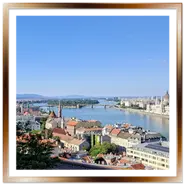 Ampia vista panoramica di Budapest dall'alto della collina di Buda. Sulla destra si staglia l'imponente Palazzo del Parlamento, al centro il fiume Danubio si estende verso l'isola Margherita e il suo ponte, mentre in primo piano spiccano i tetti colorati della città sotto un cielo azzurro e limpido.