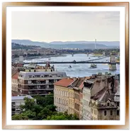 Vista panoramica dall'alto di Budapest scattata da Buda. In primo piano si stagliano i tetti degli edifici storici e alberi verdi, al centro il Ponte delle Catene attraversa il fiume Danubio animato da battelli, e sulla destra svetta la cupola del Palazzo del Parlamento con le colline sullo sfondo.