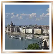 Veduta panoramica dall'alto di Buda con il Ponte delle Catene in primo piano a destra, navi da crociera sul Danubio e il Palazzo del Parlamento ungherese a Pest sullo sfondo sotto un cielo azzurro con nuvole bianche.