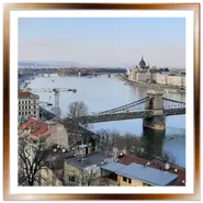 Vista panoramica dall'alto di Budapest con il Ponte delle Catene in primo piano che attraversa il fiume Danubio, il Palazzo del Parlamento ungherese sullo sfondo della sponda di Pest e i tetti della città sotto un cielo chiaro.