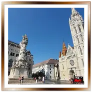 Piazza della Santissima Trinità a Budapest con la Chiesa di Mattia sulla destra, la colonna della peste al centro e il palazzo del Ministero delle Finanze sulla sinistra sotto un cielo azzurro.