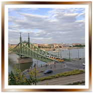 Veduta panoramica del Ponte della Libertà a Budapest in metallo verde sul Danubio, con un tipico tram giallo che lo attraversa, auto in strada e navi da crociera sullo sfondo sotto un cielo nuvoloso.
