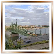 Veduta panoramica del Ponte della Libertà a Budapest in metallo verde sul Danubio, con un tipico tram giallo che lo attraversa, auto in strada e navi da crociera sullo sfondo sotto un cielo nuvoloso.
