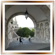 Grande arco in pietra bianca del Bastione dei Pescatori a Budapest, ornato con statue di soldati storici nelle nicchie laterali, che incornicia una scalinata all'aperto con visitatori e una torre neogotica sullo sfondo.