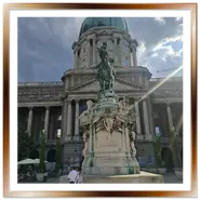 La statua equestre in bronzo di Eugenio di Savoia situata sul piazzale del Palazzo Reale nel Castello di Buda a Budapest, con la grande cupola verde della Galleria Nazionale Ungherese sullo sfondo e i raggi del sole che filtrano tra le nuvole.