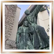 Inquadratura dal basso della statua in bronzo con patina verde di Frate Giuliano e Frate Gerardo a Budapest, con i resti di mura medievali in pietra e l'edificio moderno dell'Hilton sullo sfondo.