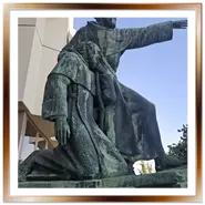 Primo piano del monumento in bronzo ai frati domenicani ed esploratori ungheresi Giuliano e Gerardo a Budapest, con l'iscrizione latina ben visibile alla base e un cielo azzurro sullo sfondo.