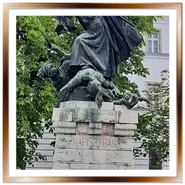 Primo piano del monumento in bronzo ai frati domenicani ed esploratori ungheresi Giuliano e Gerardo a Budapest, con l'iscrizione latina ben visibile alla base e un cielo azzurro sullo sfondo.