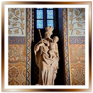 Statua in pietra della Madonna col Bambino all'interno della Chiesa di Mattia a Budapest, posizionata davanti a una vetrata colorata e circondata da pareti finemente affrescate con motivi geometrici e floreali neogotici.