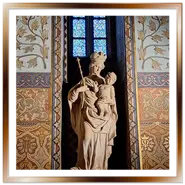 Statua in pietra della Madonna col Bambino all'interno della Chiesa di Mattia a Budapest, posizionata davanti a una vetrata colorata e circondata da pareti finemente affrescate con motivi geometrici e floreali neogotici.