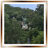 La statua in bronzo di San Gerardo con la croce alzata, situata sul pendio della collina Gellért a Budapest, circondata da fitti alberi verdi sotto un cielo parzialmente nuvoloso.