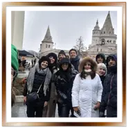 Gruppo di turisti italiani sorridenti e coperti con abiti invernali durante una visita guidata privata a Budapest, con le torri bianche del Bastione dei Pescatori sullo sfondo.