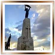 La statua della Cittadella di Budapest che si staglia contro un cielo nuvoloso al tramonto, con la bandiera ungherese visibile.