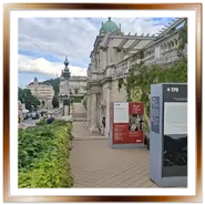 Passeggiata terrazzata del Várkert Bazár a Budapest con pannelli espositivi informativi storici, balaustre in pietra ricoperte d'edera, la statua di un leone a sinistra e la città sullo sfondo sotto un cielo nuvoloso.