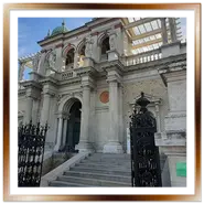 Facciata neorinascimentale del Várkert Bazár (Bazar del Giardino del Castello) a Budapest, con una scalinata in pietra, imponenti colonne, statue decorative, una cancellata in ferro battuto in primo piano e cupole verdi sotto un cielo azzurro velato.