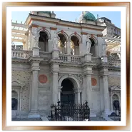Facciata monumentale del Várkert Bazár a Budapest con due grandi statue di leoni in pietra in primo piano, scalinate di accesso, un cancello in ferro battuto e il porticato del Giardino del Castello che sale verso il Palazzo Reale sullo sfondo.