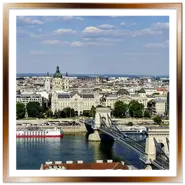 Vista panoramica dall'alto di Budapest scattata da Buda, con lo storico Ponte delle Catene che attraversa il Danubio in primo piano, la grande cupola della Basilica di Santo Stefano a Pest e grandi navi da crociera ormeggiate lungo il fiume sotto un cielo parzialmente nuvoloso.
