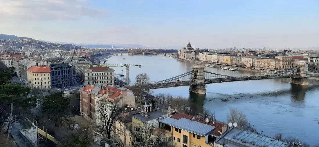 Panorama di Budapest dalla Collina del Castello – Vista su Pest Spettacolare panorama di Budapest visto dalla collina di Buda, con il Ponte delle Catene sul Danubio, il Parlamento ungherese sullo sfondo e la città di Pest sotto un cielo azzurro e nuvoloso.